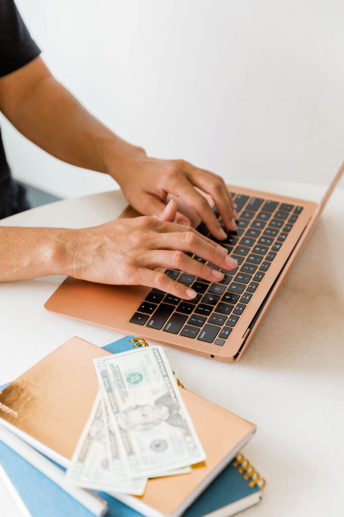 woman typing in laptop with cash on the side, counting profits